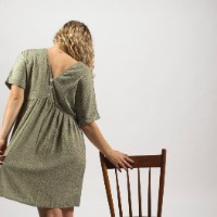 The back of a woman in a green dress with one hand on a wooden chair.